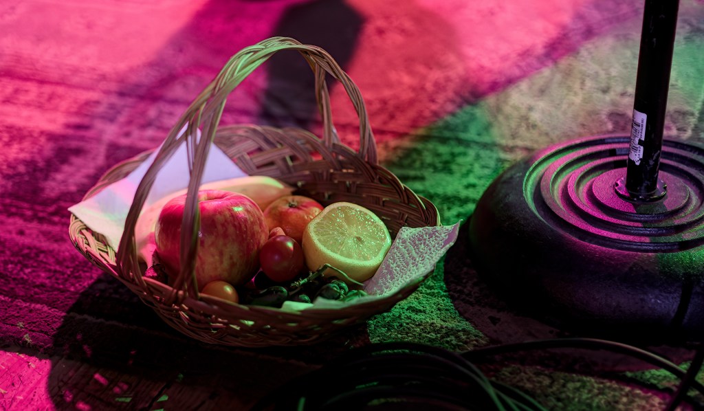 A woven basket containing various fruits, including apples, a lemon, small tomatoes, and a napkin, placed on a colorful textured surface with a microphone stand nearby.