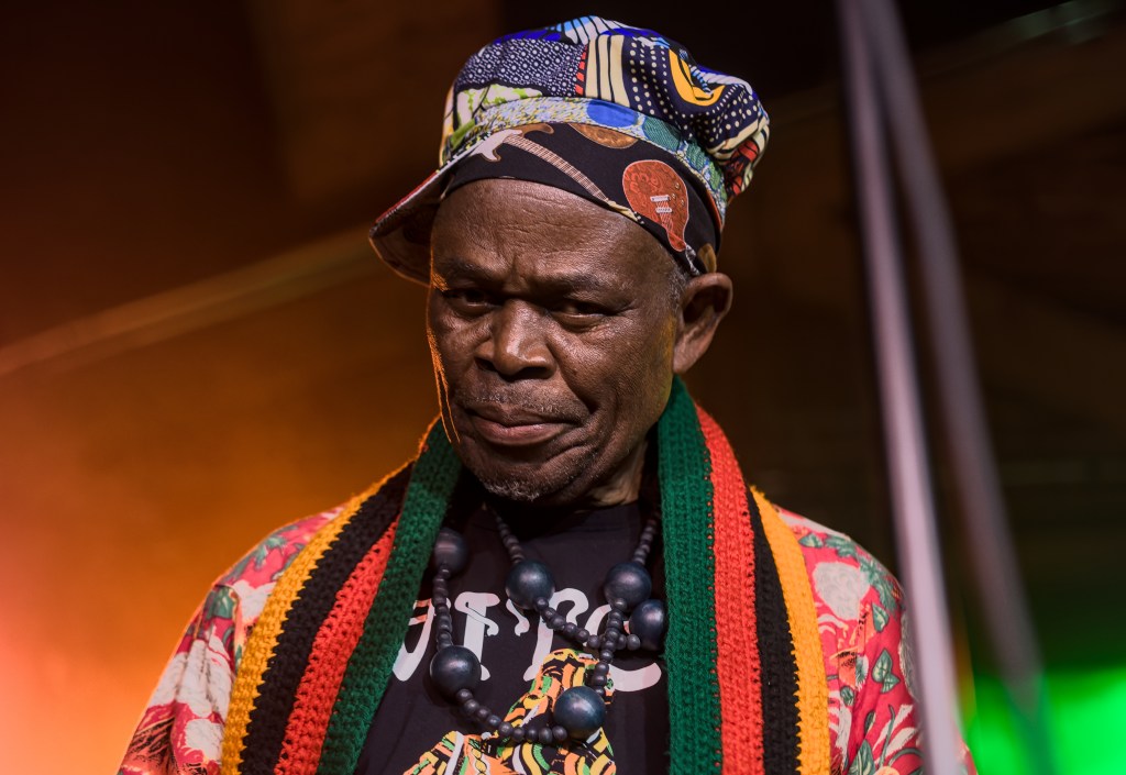 A close-up portrait of an older man wearing a colorful hat and a patterned shirt, with a serious expression and layered beaded necklaces, set against a softly lit background.