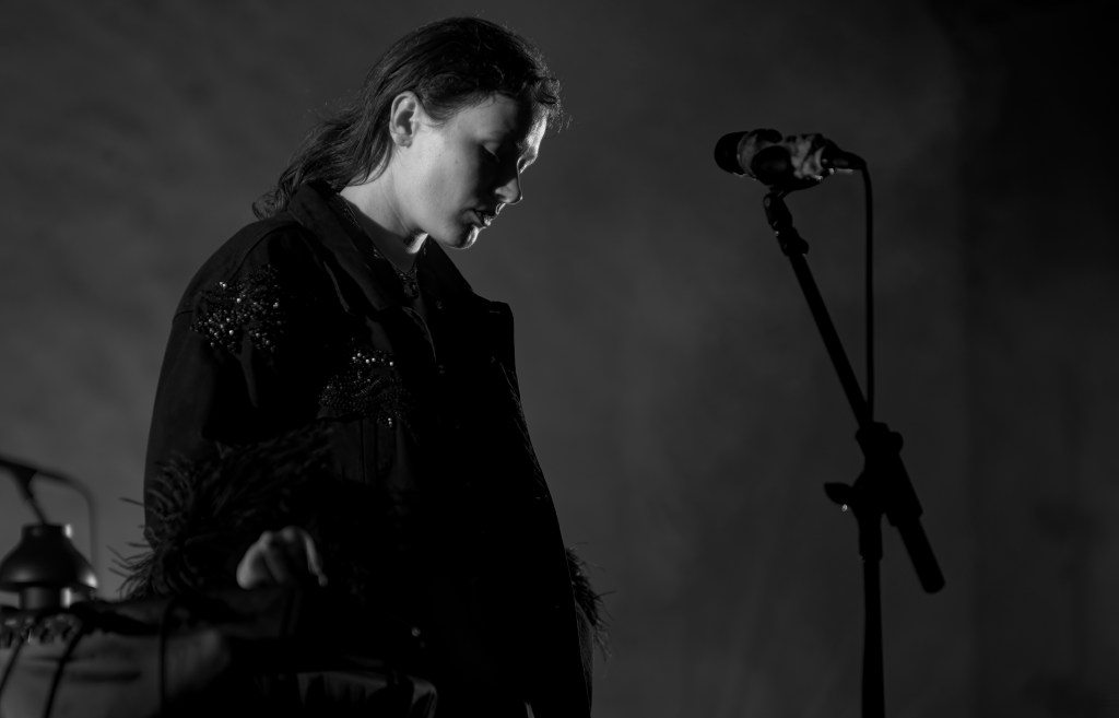 A performer in a black jacket with decorative details, standing thoughtfully beside a microphone in a dimly lit setting.