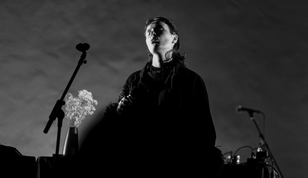 A musician performing on stage in black and white, with a microphone and a vase of flowers visible in the foreground.