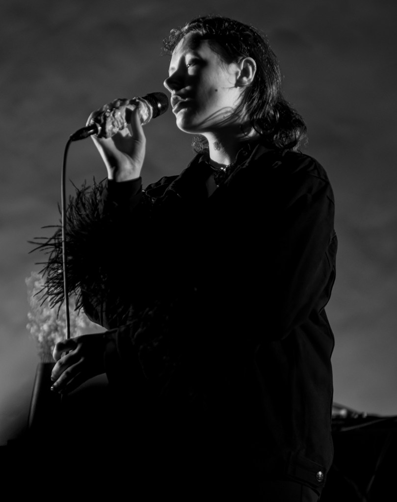 A vocalist performing on stage with a microphone, captured in black and white. The performer has long hair and wears a dark outfit adorned with feathers. Their expression is focused and emotive.