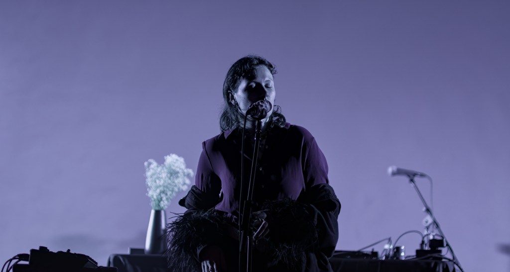 A female performer singing into a microphone on stage, with a minimalist setup including a flower arrangement and electronic equipment, illuminated by soft purple lighting.