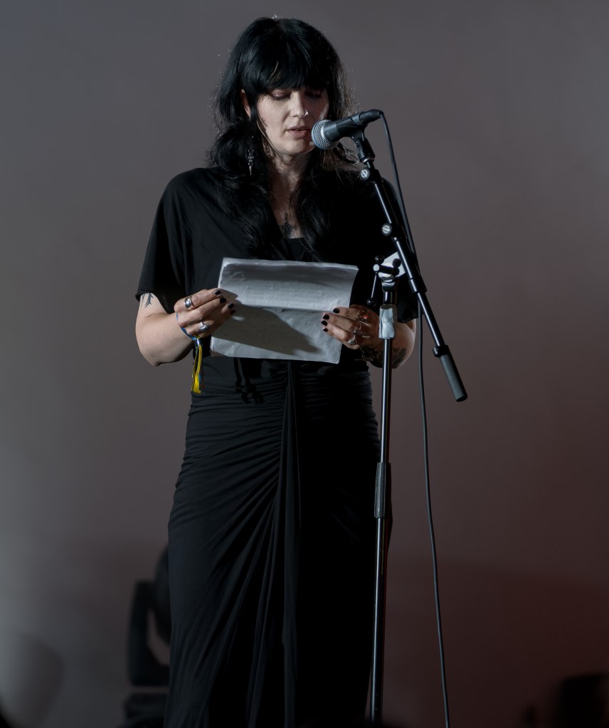 A woman with long black hair reads from a piece of paper while standing at a microphone, wearing a black dress and accessories.