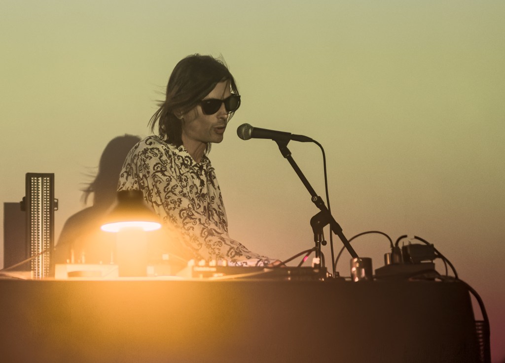 A musician performing on stage at sunset, wearing sunglasses and a patterned shirt, with a microphone and various electronic equipment on the table.