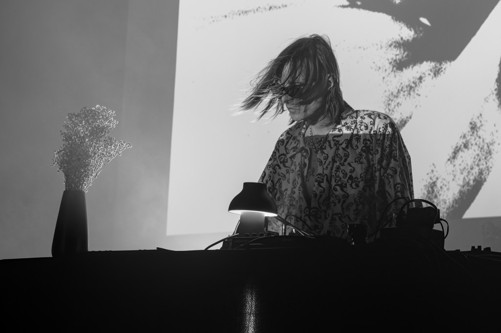 A DJ performing on stage with long hair blowing in the air, wearing a patterned top, with a vase of flowers and equipment on the table. In the background, a projected abstract image.
