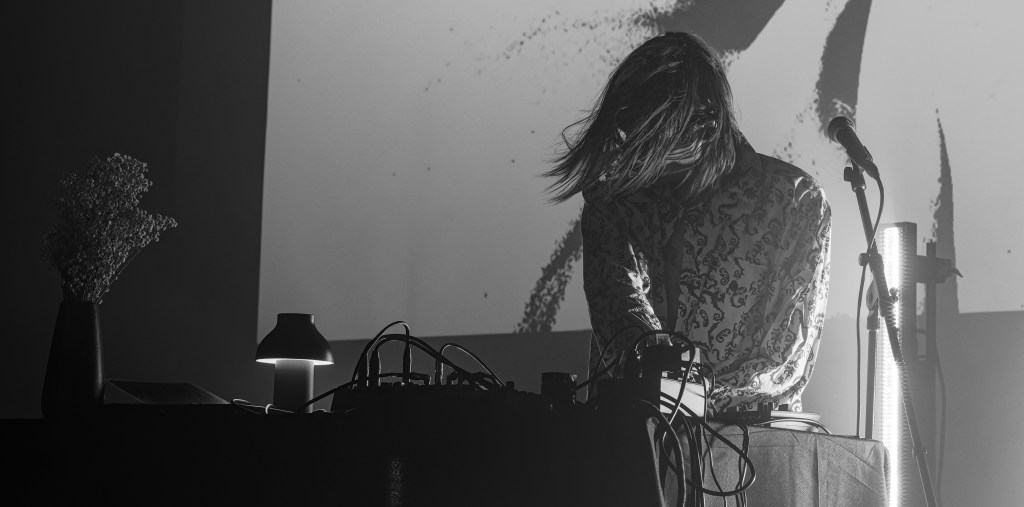 A musician performing on stage with long hair, wearing a patterned shirt, surrounded by electronic equipment and a microphone, in a dimly lit environment with a decorative vase and lamp.