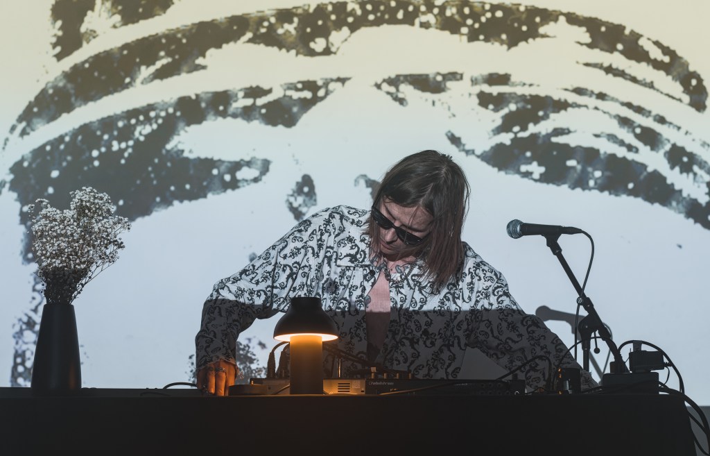 A musician performing at a live event, with a projector screen displaying abstract visuals in the background. The performer, wearing sunglasses and a patterned shirt, is focused on a turntable, amidst a table decorated with a lamp and a small bouquet of flowers.