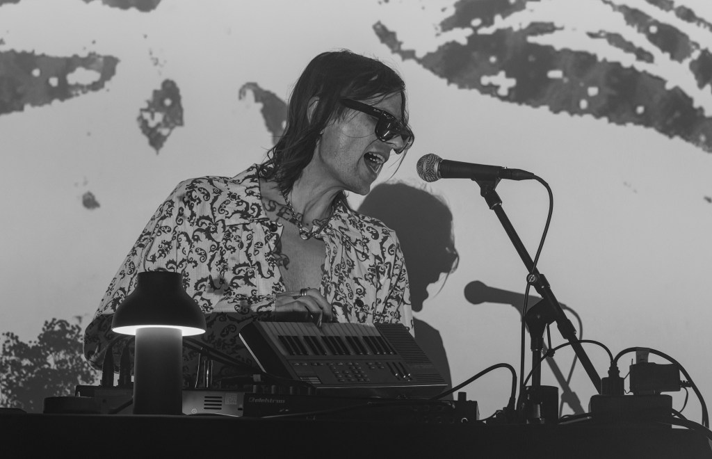 A musician performing on stage, wearing sunglasses and a patterned shirt, passionately singing into a microphone with sound equipment and a lamp on the table.