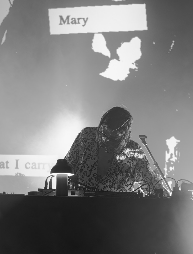 A DJ performing live on stage, wearing a patterned shirt, with a microphone and equipment in front of them, while a projected screen in the background displays text.