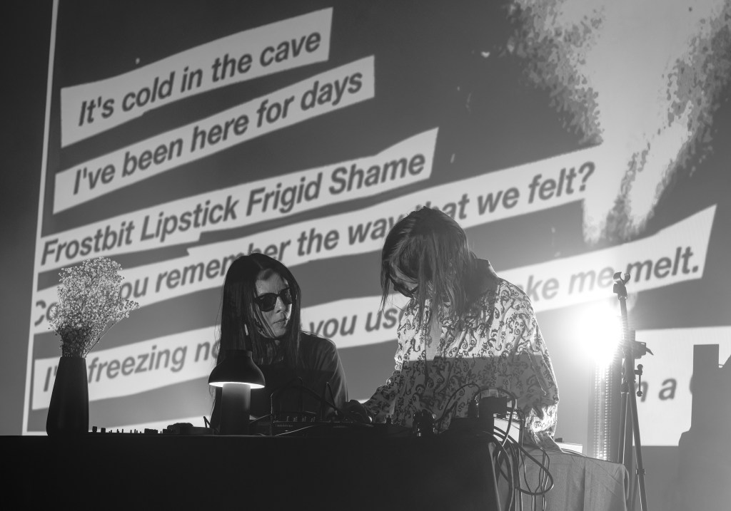 A black and white image of two musicians performing on stage, focused on their electronic instruments, with text projections in the background.