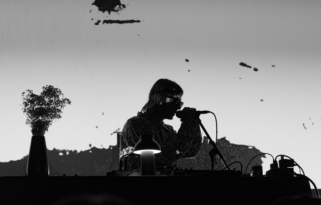 A silhouette of a musician performing with a microphone, surrounded by a sound system and a flower vase, against a stark, textured backdrop.