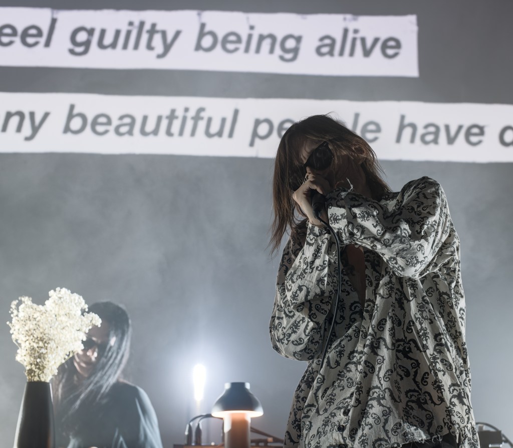 A singer performing on stage with a microphone, wearing a patterned shirt, while a blurred figure stands behind them. Projected text in the background includes poetic phrases.