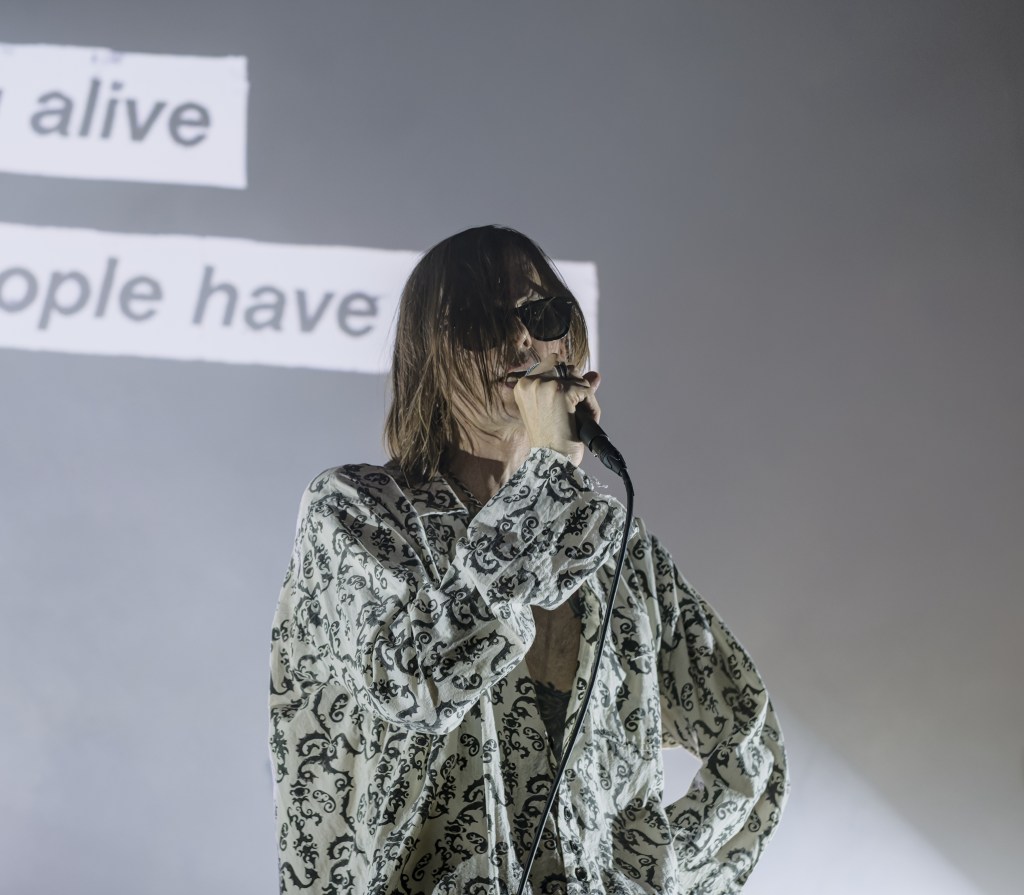 A musician performing on stage, wearing sunglasses and a patterned shirt, singing into a microphone with a blurred projection in the background featuring text.