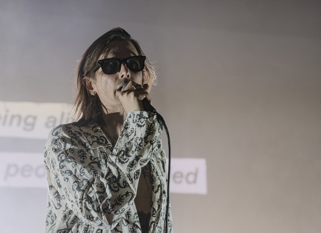 A singer wearing sunglasses and a patterned shirt performs on stage, microphone in hand, with a blurred background displaying text.