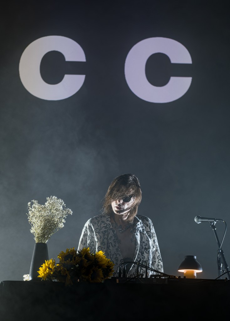 A musician performing on stage in front of large illuminated letters 'C C', surrounded by flowers and equipment.