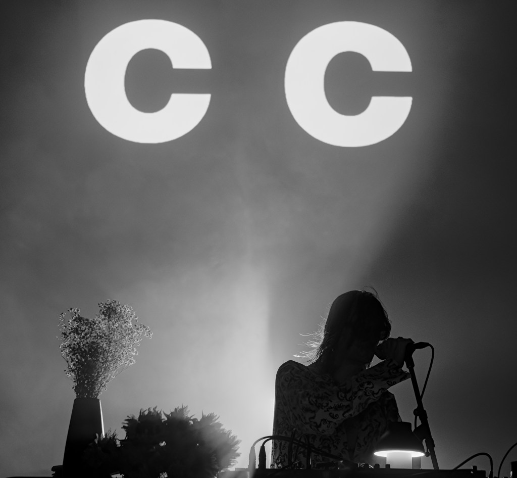 A silhouette of a performer singing into a microphone on stage, with large letters 'CC' illuminated in the background. A flower arrangement and electronic equipment are visible in the foreground, set against dramatic lighting.