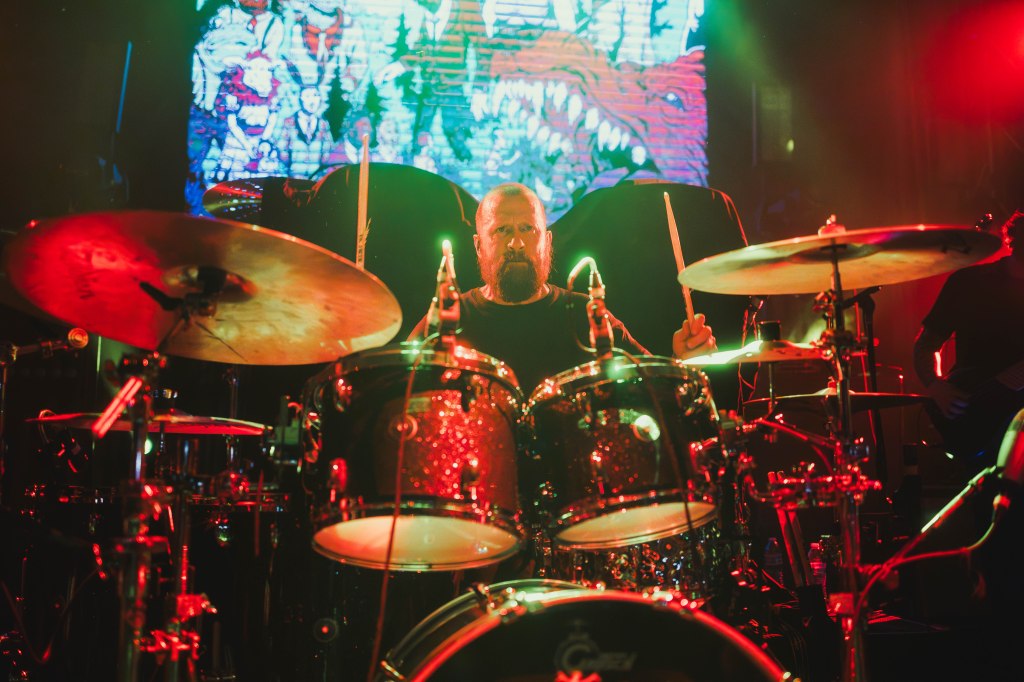 A drummer performing on stage with colorful lights in the background, showcasing a dynamic live music atmosphere.