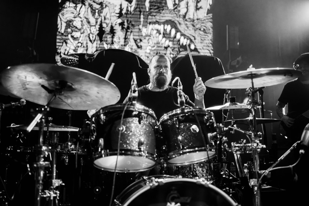 A drummer playing passionately on stage, with a focus on his facial expression and drum set. The background features vibrant visuals and lighting effects, creating an energetic atmosphere.
