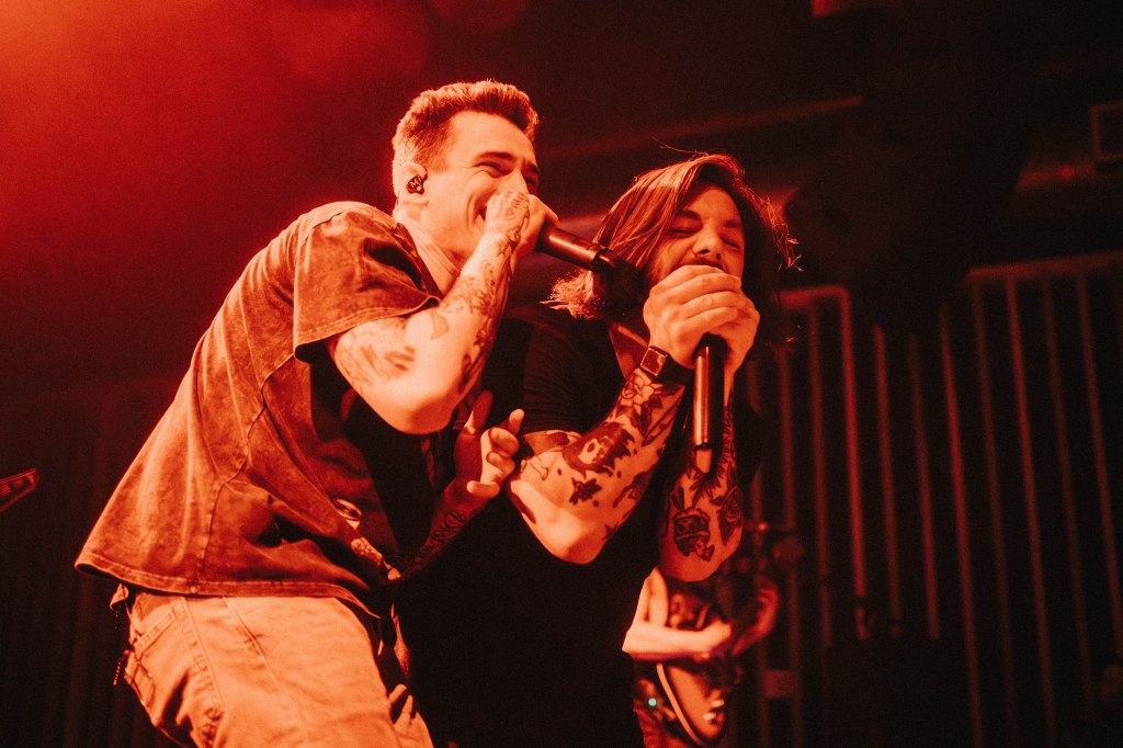 Two musicians performing on stage, one singing into a microphone while the other leans in closely with a joyful expression. The lighting is warm with an orange hue, enhancing the lively atmosphere of the concert.