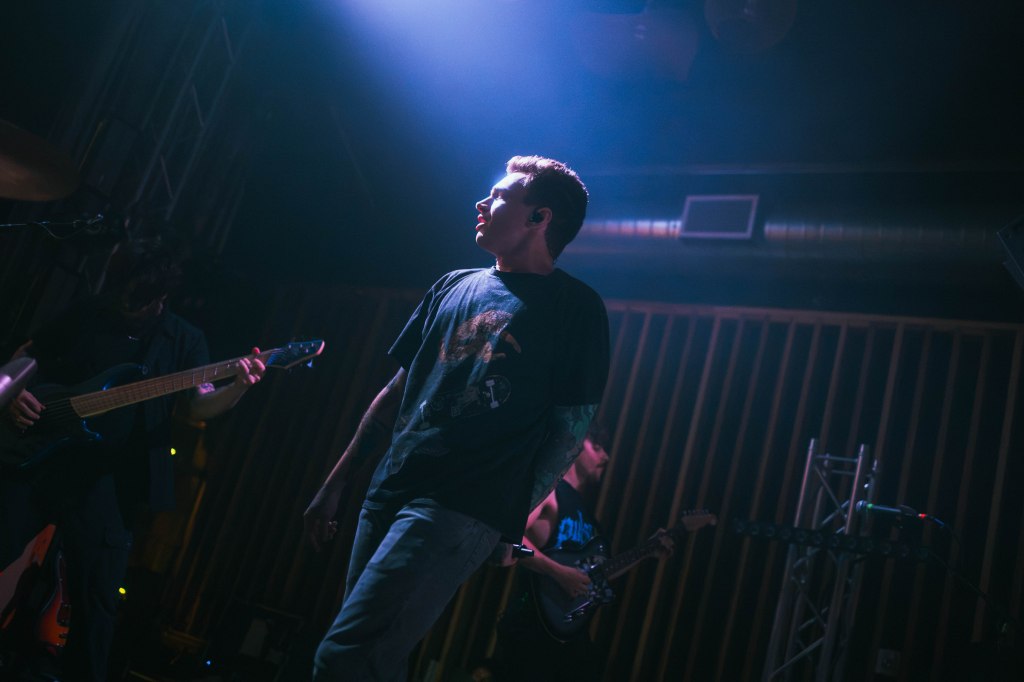 A musician performing on stage with dramatic lighting, wearing a black t-shirt and jeans, while holding a microphone.