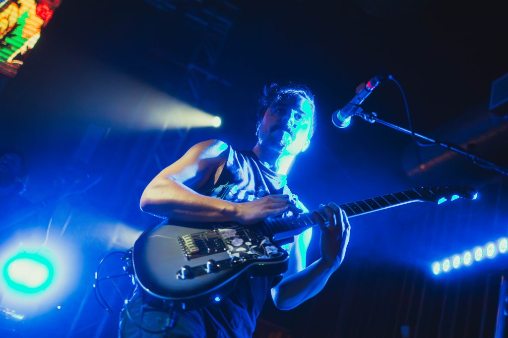 A musician performing on stage with a guitar, surrounded by blue lighting and an energetic atmosphere.