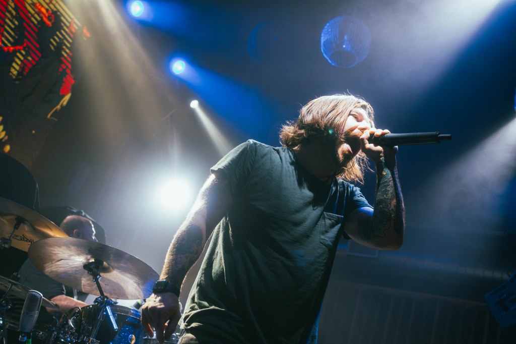 A male vocalist passionately performing on stage with a microphone, illuminated by colorful lights, while a drummer is visible in the background.
