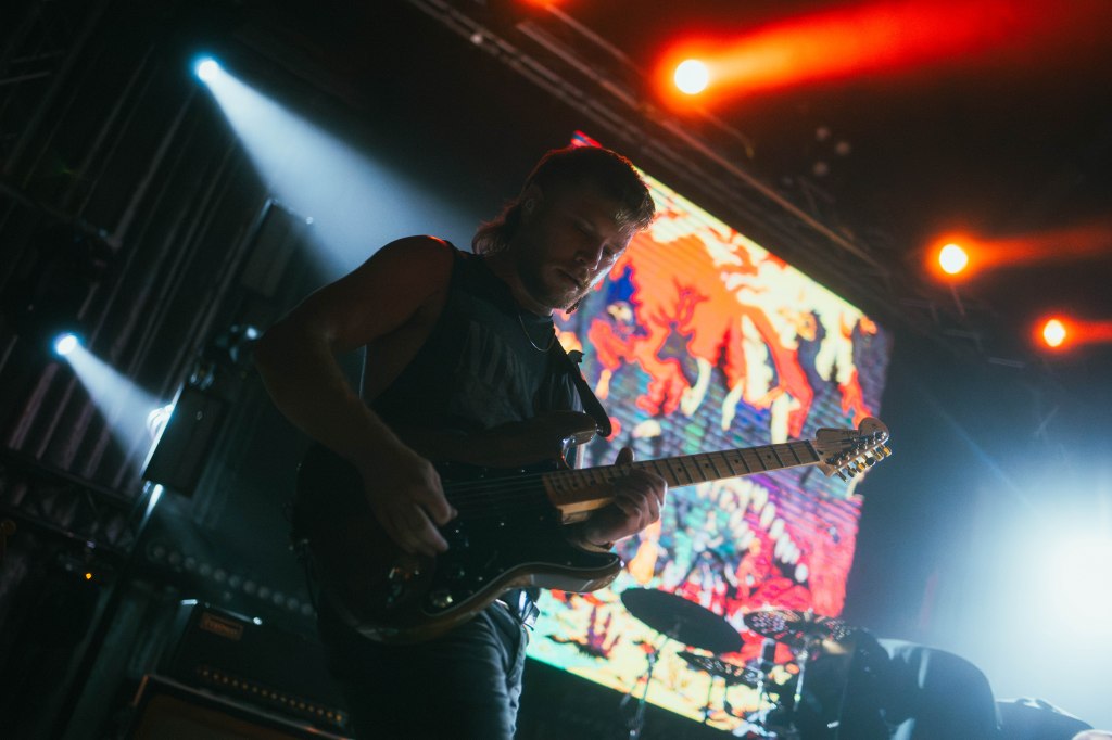 A guitarist performing on stage under colorful lighting with a vibrant backdrop.