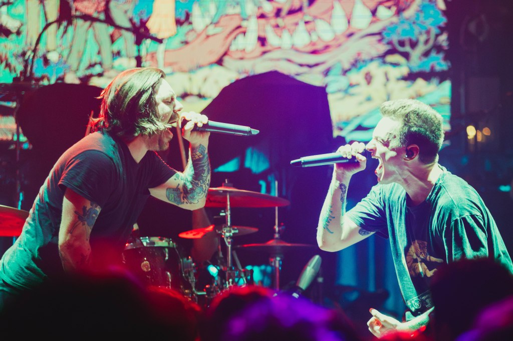 Two male singers performing on stage with microphones, passionately engaging with the audience, against a colorful backdrop.