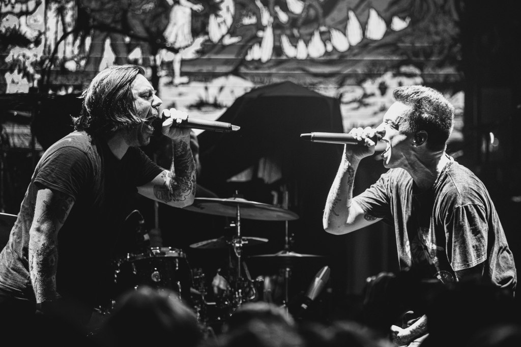Two male singers passionately performing on stage with microphones in hand, surrounded by a dramatic backdrop and a drum set.