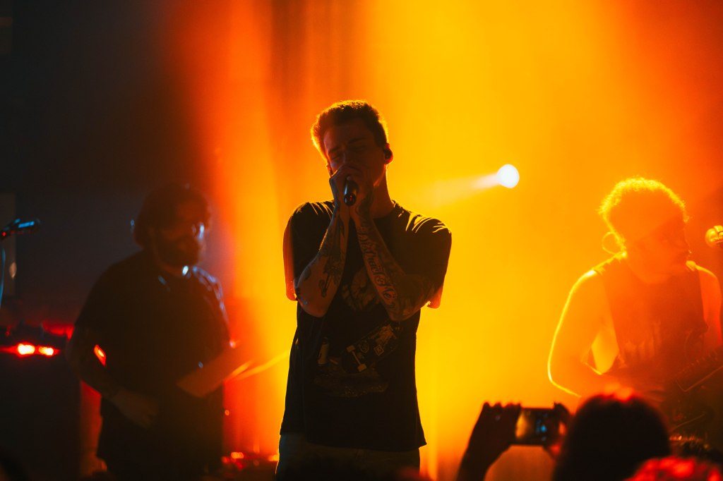 A male singer passionately performing on stage in front of an audience, surrounded by warm orange lights, with a guitarist and a band member visible in the background.