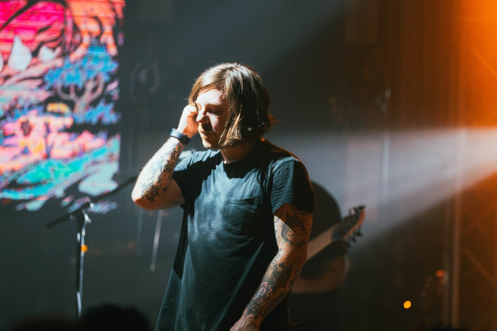 A musician with tattoos performing on stage, looking thoughtful under dramatic lighting, with a colorful backdrop behind him.
