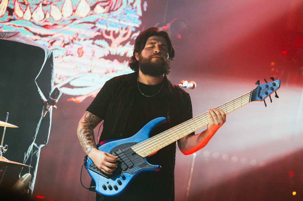 A male musician playing a blue six-string bass guitar on stage, surrounded by colorful lighting and a dynamic backdrop.