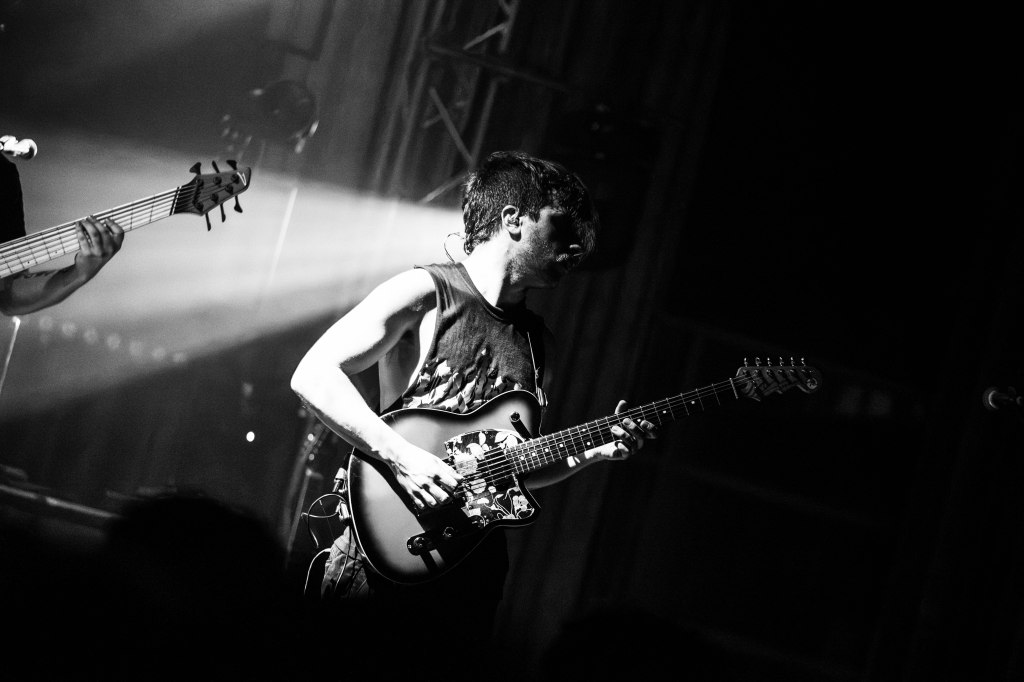 A musician performing on stage with an electric guitar, illuminated by stage lights in a black and white setting.