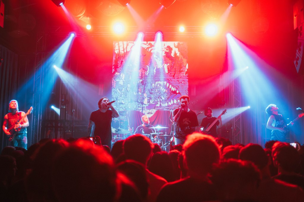A live music performance featuring a band with multiple musicians on stage, vibrant lighting, and an enthusiastic audience in front.