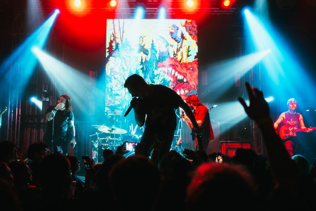 A lively concert scene featuring a band on stage with vibrant lighting and a colorful backdrop. The lead singer engages with the crowd while band members play instruments.
