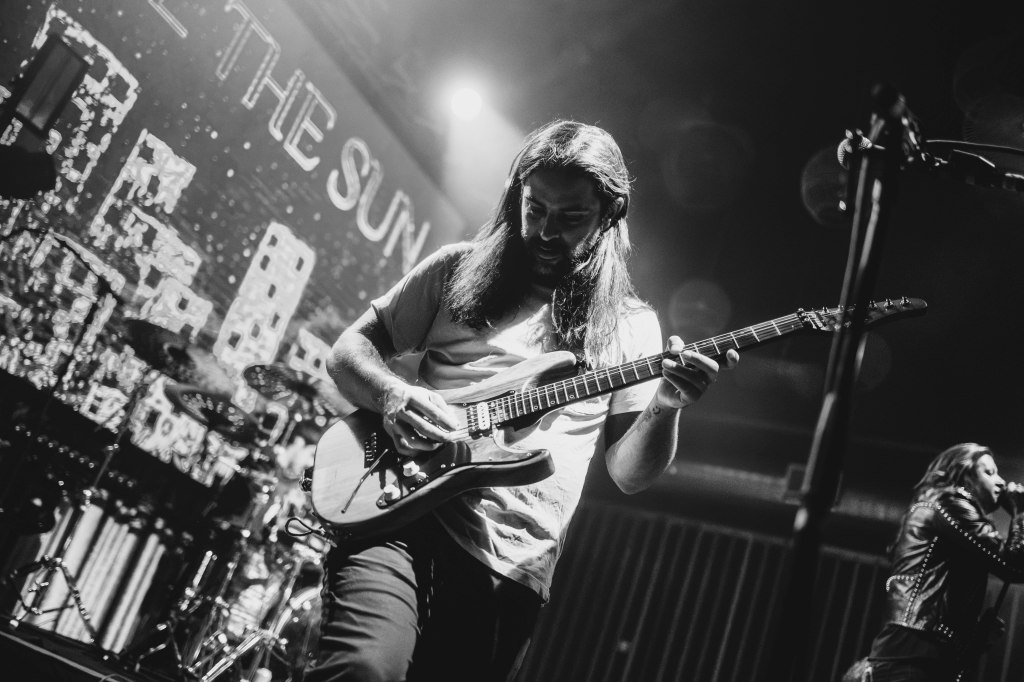 A guitarist performing on stage, focusing on playing his guitar, with a drummer and singer visible in the background. The image is in black and white, capturing the energy of a live music performance.