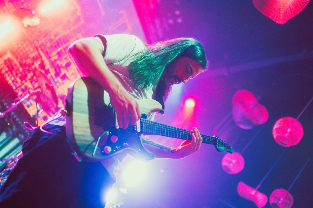 A musician with long hair playing an electric guitar on stage, surrounded by colorful lights and hanging lanterns.