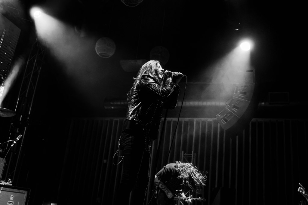 A performer singing into a microphone on stage, wearing a black leather jacket, with dramatic lighting and shadows creating a dynamic atmosphere.