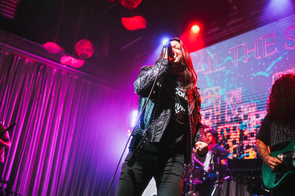 A lead singer performing on stage with a microphone, wearing a leather jacket and a graphic t-shirt, surrounded by colorful stage lights. A band is visible in the background.