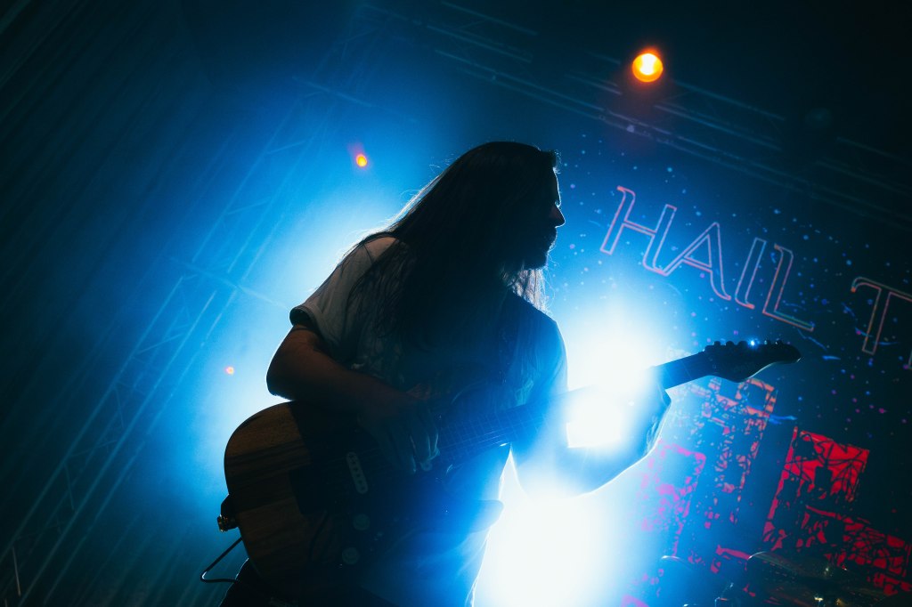 Silhouette of a guitarist on stage, illuminated by bright blue and white lights with a 'HAIL THE' banner in the background.