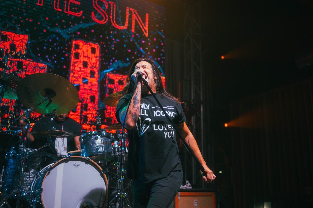 A lead singer performing on stage in a black t-shirt, passionately engaging with the audience while a drummer plays behind them, with vibrant stage lights and colorful visuals in the background.