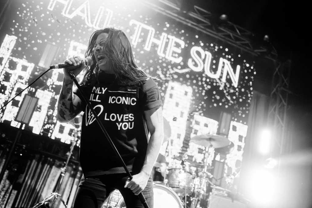 A musician performing on stage, passionately singing into a microphone, wearing a black t-shirt with the text 'ONLY ICONIC LOVES YOU.' The background features bright lights and a banner displaying the name 'HAIL THE SUN.'