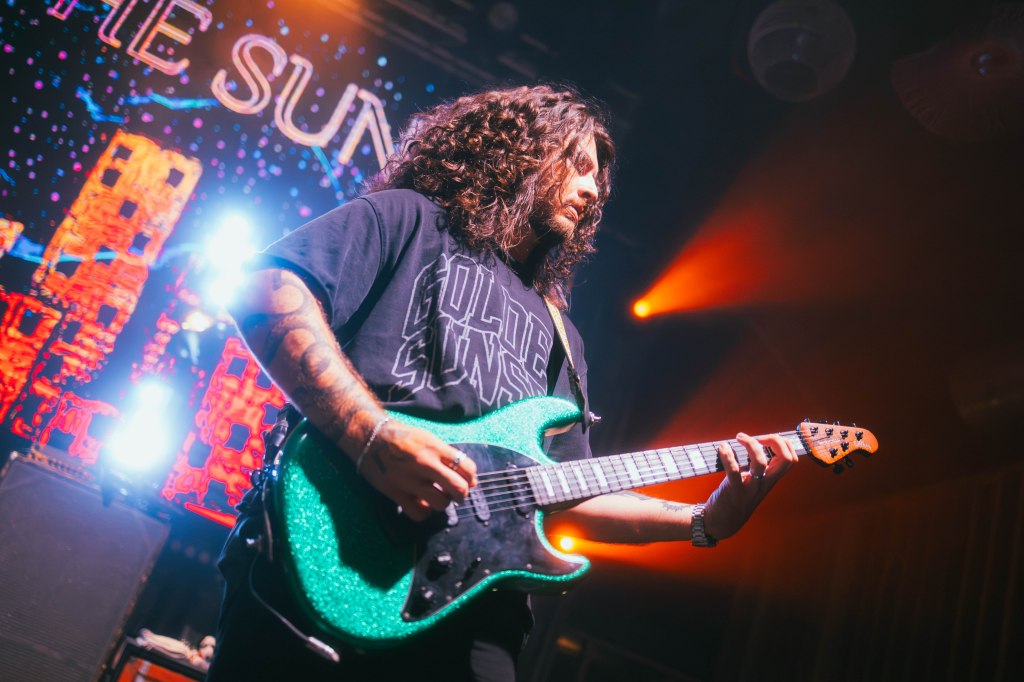 A musician playing a green electric guitar on stage, with colorful lights and a digital backdrop featuring the words 'GOLDEN SUN'.
