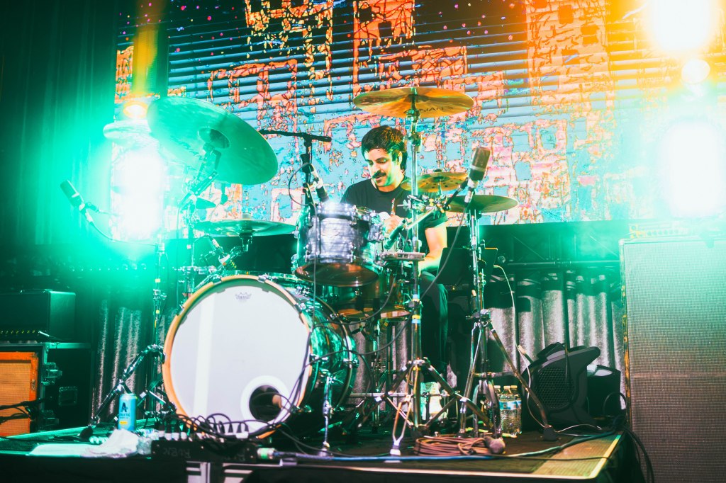 A drummer performing on stage with colorful lighting and a vibrant background showcasing sound equipment.