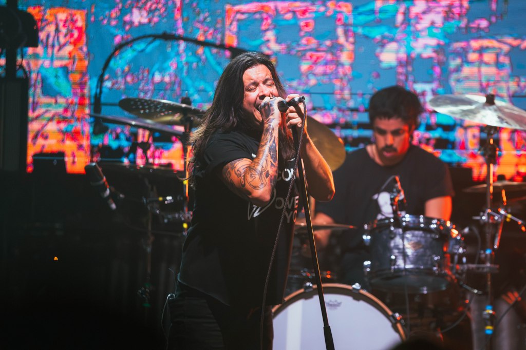 A singer with long hair passionately performing on stage while holding a microphone, with a drummer visible in the background and colorful lights illuminating the scene.