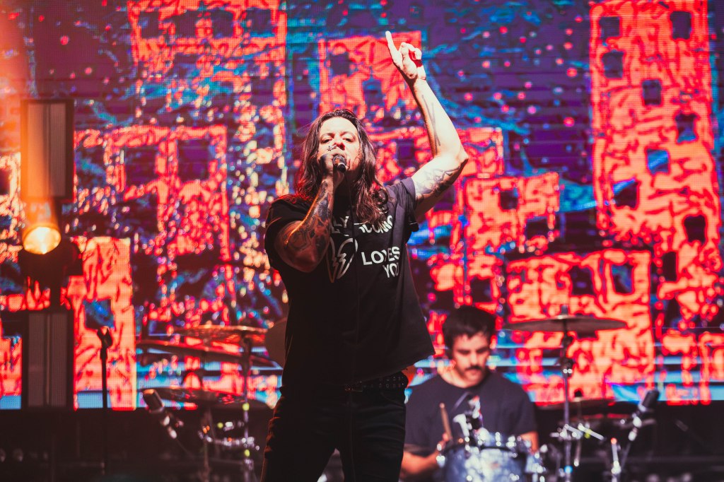 A musician performing on stage with a raised hand, wearing a black t-shirt with a graphic design, set against a vibrant and colorful background with a band playing behind.