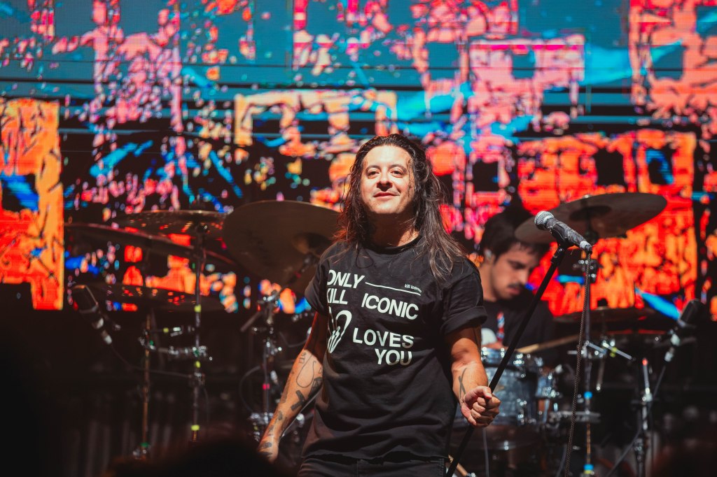 A singer on stage wearing a black t-shirt with the text 'ONLY ICONIC KILL LOVES YOU,' smiling with long hair, in front of a vibrant, multicolored background and a drummer in the background.