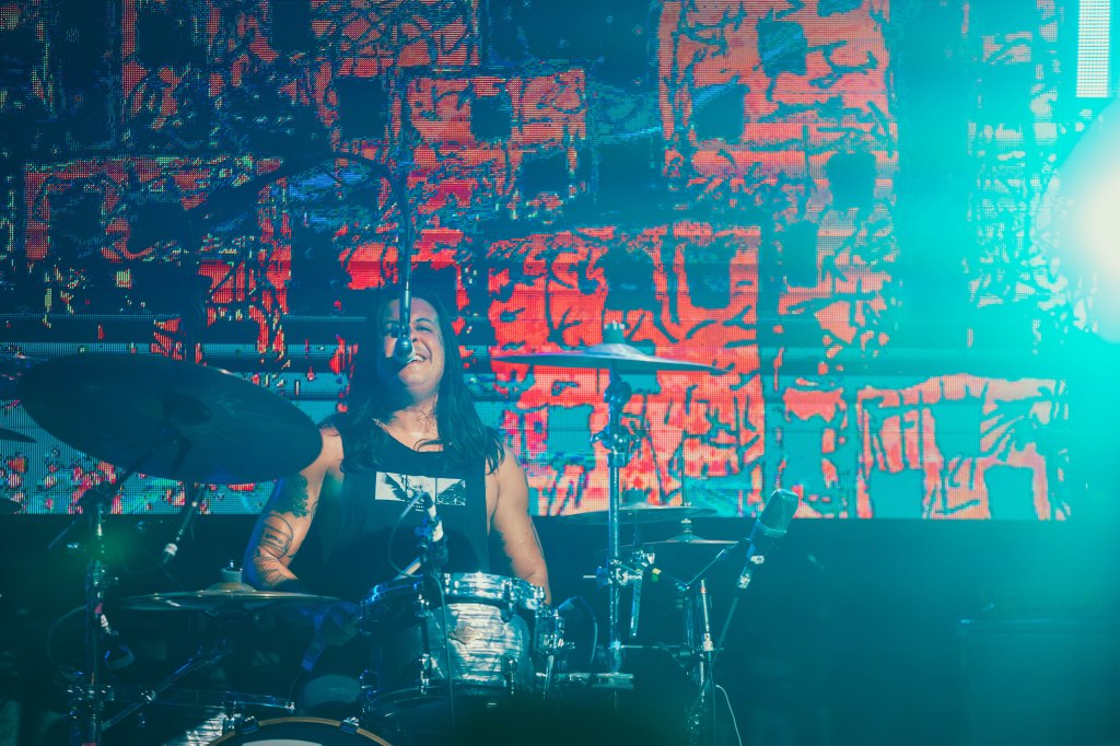 A drummer performing on stage, smiling and engaging with the audience, with colorful lights and a vibrant background.