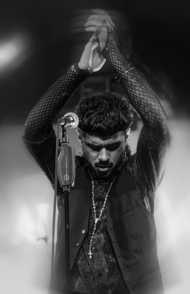 A performer with dark hair and a stylish outfit, raising hands in a dramatic pose, standing in front of a microphone, captured in black and white.