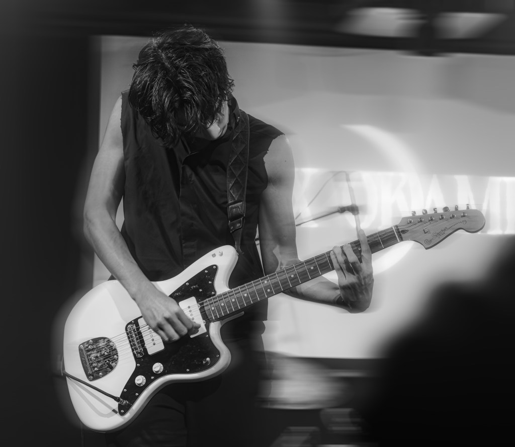 A musician playing an electric guitar on stage, captured in motion, with a focus on the guitar and hands, set against a blurred background.